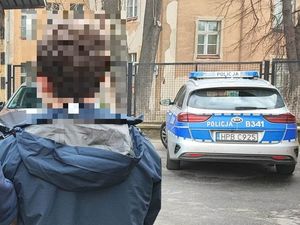 Poszukiwany mężczyzna stoi tyłem do zdjęcia ubrany w niebieską kurtkę. Zdjęcie wykonane na parkingu policyjnym w tle oznakowany radiowóz policyjny.