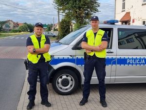 Dwóch policjantów ubranych w mundury służbowe oraz kamizelki odblaskowe stoją na tle oznakowanego radiowozu policyjnego typu bus