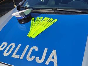 Czapka policjanta ruchu drogowego z białym otokiem oraz odblaski w formie linijek leżą na masce radiowozu policyjnego. Znajduje się tam również napis Policja.