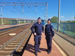 Policjant oraz policjantka w mundurach ćwiczebnych typu moro stoją na peronie kolejowym. Jest ładna słoneczna pogoda. Oboje uśmiechają się.