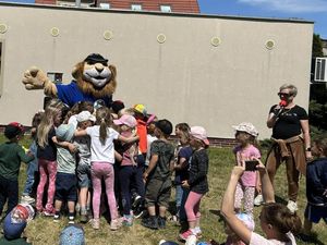 Na placu zabaw w słoneczną pogodę grupka dzieci z przedszkola tuli się do maskotki dolnośląskiej Policji Komisarza Lwa.