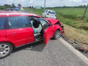 Czerwony samochód osobowy uszkodzony w wyniku wypadku. Uszkodzeniu uległ przód oraz prawy bok samochodu. Pogoda jest słoneczna, pojazd stoi w poprzek drogi.