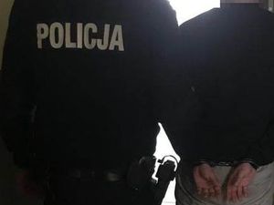 Policjant w mundurze na plecach ma napis Policja wraz z mężczyzną, który ma założone kajdanki na ręce trzymane z tyłu.