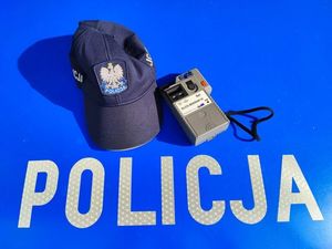 Na masce oznakowanego radiowozu z napisem Policja leży czapka policyjna z daszkiem oraz urządzenie do badania stanu trzeźwości.