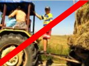 Dziecko stoi na łączeniu pomiędzy traktorem a przyczepą. Cale zdjęcie przekreślone jest czerwoną linią.