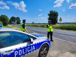 Policjanci przy drodze wykonują pomiar prędkości, jeden trzyma urządzenia drugi policjant stoi przy oznakowanym radiowozie