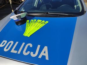 Na masce oznakowanego radiowozu policyjnego z napisem Policja położona jest czapka policjanta z ruchu drogowego z białym otokiem oraz opaski odblaskowe.