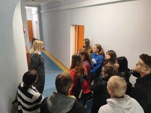 Na korytarzu komisariatu policjanta przedstawia pracę Ogniwa do spraw Kryminalnych, przed nią w grupie stoją uczniowie.