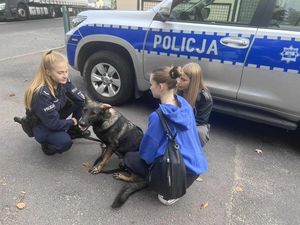 Policjantka, przewodniczka psa służbowego kuca obok swojego czworonożnego funkcjonariusza, przed nią kuca jedna z uczennic. W tle znajduje się oznakowany radiowóz policyjny.