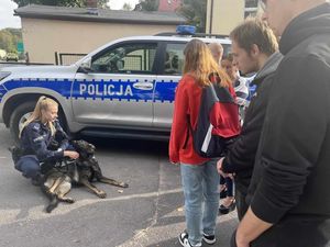 Policjantka z psem służbowym kuca, a przed nią stoi grupa uczniów. W tle oznakowany radiowóz policyjny.