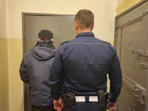 Policjant w mundurze służbowym oraz mężczyzna zatrzymany. Obaj stoją tyłem do zdjęcia. Zdjęcie wykonywane na terenie komendy.