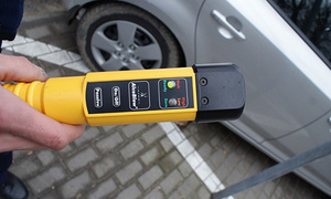 Urządzenie do badania stanu trzeźwości trzymane w ręce na tle samochodu. Urządzenie jest koloru żółtego.