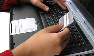Laptop dwie dłonie oparte na klawiaturze. W jednej dłoni osoba trzyma kartę bankomatową.