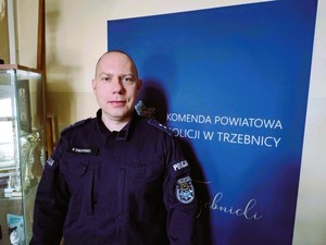 Policjant umundurowany stoi obok banera Komendy Powiatowej Policji w Trzebnicy