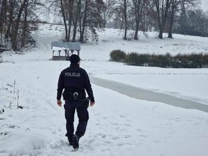 Policjant w mundurze służbowym kontroluje zbiorniki wodne oraz pola gdzie może dochodzić do organizacji kuligów. W miejscu gdzie znajduje się policjant leży śnieg a woda jest zamarznięta.
