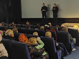 Policjanci prowadzą szkolenie dla wolontariuszy. Spotkanie odbywa się w sali kinowej. Policjanci stoją na scenie, wolontariusze siedzą w fotelach.