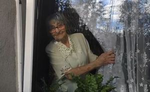 Starsza kobieta odchyla firany w oknie i wygląda za zaproszonymi gośćmi. Kobieta uśmiecha się.