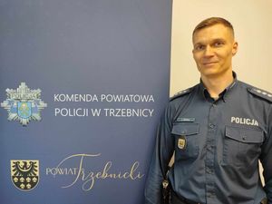 Policjant w mundurze służbowym stoi obok baneru Komendy Powiatowej Policji w Trzebnicy