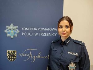Policjantka w mundurze służbowym stoi obok baneru Komendy Powiatowej Policji w Trzebnicy, Zdjęcie zrobione w pomieszczeniu, policjantka uśmiecha się.