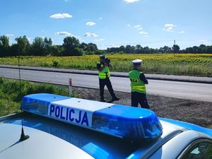 Dwóch policjantów ruchu drogowego w mundurach służbowych oraz kamizelkach odblaskowych stoją przy oznakowanym radiowozie przy ulicy. Jeden z policjantów dokonuje pomiaru prędkości trzymając w rękach radar. Pogoda jest słoneczna.