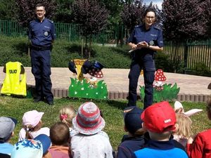 Zdjęcie przedstawia policjantkę oraz policjanta, którzy prowadzą spotkanie z przedszkolakami. Dzieci siedzą na trawie.