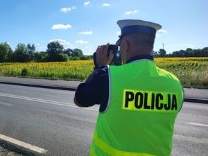 Zdjęcie przedstawia policjanta stojącego tyłem w kamizelce odblaskowej z napisem Policji, który dokonuje pomiaru prędkości