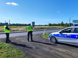 Zdjęcie przedstawia dwóch policjantów stojących tyłem ubranych w kamizelki odblaskowe z napisem policja dokonujących pomiaru prędkości przy drodze