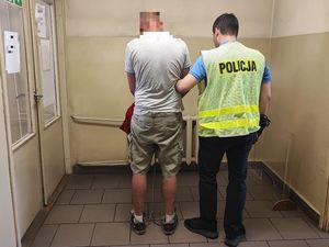 Zatrzymany mężczyzna stoi tyłem do zdjęcia wraz z policjantem kryminalny ubranym w odblaskową kamizelkę z napisem Policja