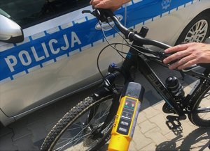 Na tle radiowozu i napisu policja widoczny jest rower oraz urządzenie do pomiaru alkoholu w organiźmie