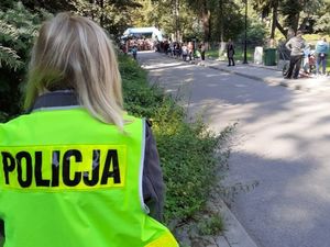 Policjantka ubrana w kamizelkę odblaskową z napisem Policja stoi tyłem do zdjęcia, a w tle rozpoczynający się wyścig