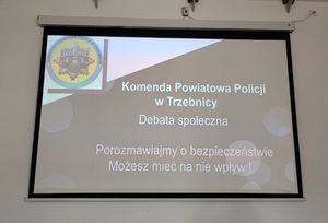 Na telebimie wyświetlona jest prezentacja, na której znajduje się Znak Komendy Powiatowej Policji w Trzebnicy oraz napisane jest Komenda Powiatowa Policji w Trzebnicy, Porozmawiajmy o bezpieczeństwie - możesz mieć na nie wpływ