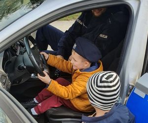 Chłopiec ma założoną czapkę policyjną na głowie i siedzi za kierownica radiowozu, obok siedzi policjant