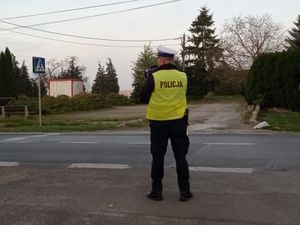 Policjant stoi przy drodze na której znajduje się przejście dla pieszych. Ubrany w mundur służbowy oraz kamizelkę odblaskową