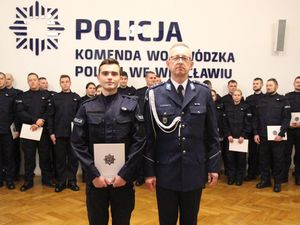 Nowo przyjęty policjant stoi obok Komendanta Powiatowego Policji w Trzebnicy. Policjant ubrany w mundur ćwiczebny, natomiast komendant w mundur galowy. Z tyłu stoją przyjęci funkcjonariusze, na ścianie napis Komenda Wojewódzka Policji we Wrocławiu