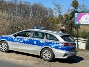 Oznakowany radiowóz policyjny, za nim stoi policjant, który będzie kontrolował ogródki działkowe