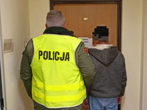 Policjant stoi w odblaskowej kamizelce z napisem Policja obok niego stoi mężczyzna poszukiwany. Stoją tyłem do zdjęcia