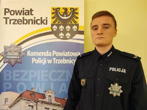 Dzielnicowy Szymon Stencel ubrany w mundur służbowy stoi obok banneru Komendy Powiatowej Policji w Trzebnicy