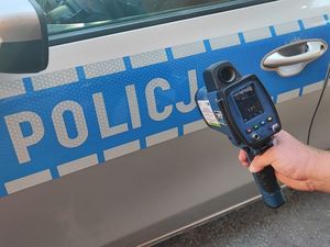 Miernik prędkości trzymany w ręce, w tle napis policja znajdujący się na oznakowanym radiowozie policyjnym