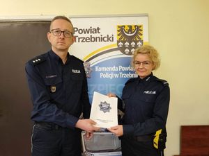 Wręczenie rozkazu przez Komendanta Powiatowego Policji w Trzebnicy nowej dzielnicowej. Wręczenie odbywa się w budynku jednostki. Policjanci ubrani w mundury służbowe, granatowe. W ręce trzymają okładkę, w której znajduje się rozkaz.