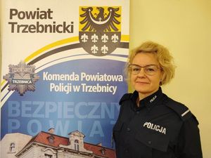 Dzielnicowa miasta Trzebnica starszy Posterunkowy Katarzyna Olechowska stoi na tle baneru Komendy Powiatowej Policji w Trzebnicy. ubrana w koszulę służbową koloru granatowego, na twarzy ma założone okulary.