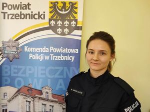 Policjantka ubrana w mundur ćwiczebny stoi przy banerze Komendy Powiatowej Policji w Trzebnicy, na którym znajduje się również logo Starostwa Powiatowego Policji w Trzebnicy