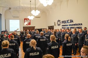 Nowo przyjęci policjanci stoją w szeregu tyłem do zdjęcia, ubrani są w mundur ćwiczebny.  Wszyscy znajdują się na auli Komendy Wojewódzkiej Policji we Wrocławiu.