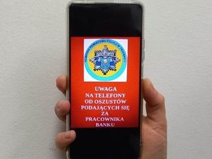 Telefon typu smartfon trzymany w ręce. Na ekranie znajduje się logo Komendy Powiatowej Policji w Trzebnicy. Pod nim znajduje się napis wielkimi literami UWAGA NA OSZUSTÓW PODAJĄCYCH SIE ZA PRACOWNIKA BANKU