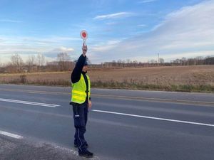 Policjant ruchu drogowego stoi przy drodze ubrany w mundur granatowy oraz odblaskową kamizelkę. Ma uniesioną rękę, w której trzyma tak zwanego &quot;lizaka&quot; do zatrzymania pojazdu.