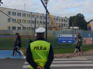 Zdjęcie przedstawia policjanta w odblaskowej kamizelce oraz czapce ruchu drogowego stojącego tyłem do zdjęcia. Policjant stoi w okolicy przejścia dla pieszych. W tle szkoła podstawowa.