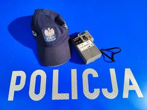 Zdjęcie przedstawia wycinek maski radiowozu oznakowanego, na którym widać napis POLICJA oraz czapkę policyjną z daszkiem oraz urządzenie do sprawdzania stanu trzeźwości.