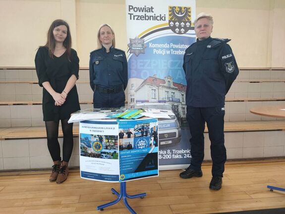 Zdjęcie przedstawia dwie policjantki oraz pracownika cywilnego trzebnickiej komendy. Przed nimi stoi stolik z ulotkami oraz gazetami policyjnymi. Za nimi znajduje się baner trzebnickiej Policji.