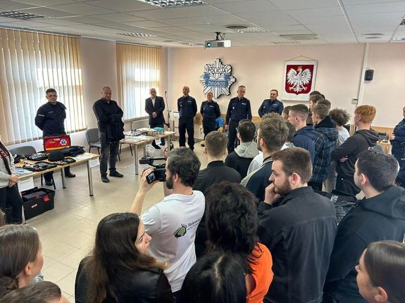 Na sali odpraw znajduje się młodzież, w oddali Komendant Powiatowy Policji w Trzebnicy wygłasza przemówienie
