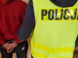 Mężczyzna z założonymi kajdankami na ręce trzymane z tyłu, widoczny jego tułów obok niego stoi policjant w kamizelce odblaskowej z napisem Policja.