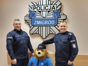 Policjanci z Komisariatu Policji w Żmigrodzie stoją obok siebie w mundurach ćwiczebnych. Uśmiechają się pomiędzy nimi stoi maskotka Komisarza Lwa, nad ich głowami znajduje się gwiazda policyjna z napisem Policja Żmigród